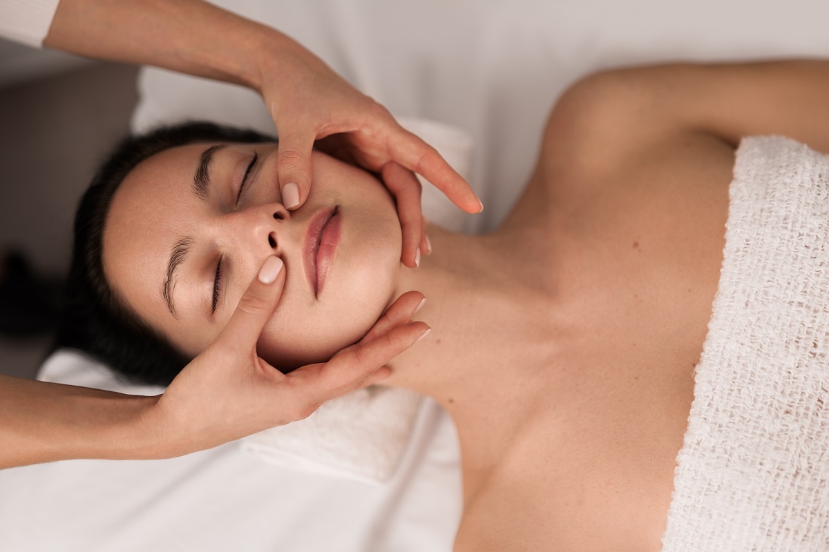 Crop masseuse massaging face of client From above massage therapist rubbing cheeks of relaxed woman during spa session in salon